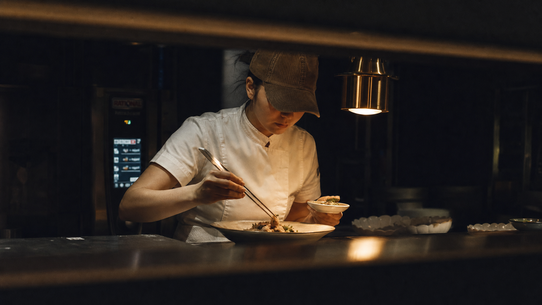 female chef at a pass in a fine-dining open kitchen, caught mid-plating